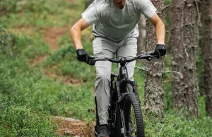 Cyklist i röd hjälm och röd tröja cyklar på muddig stig med elcykel genom uppodlat terräng, andra cyklister suddigt i bakgrunden.