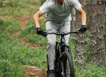 Cyklist i röd hjälm och röd tröja cyklar på muddig stig med elcykel genom uppodlat terräng, andra cyklister suddigt i bakgrunden.